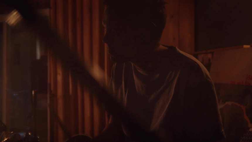 A man sitting in a room playing drums with drumsticks with a wall in the background