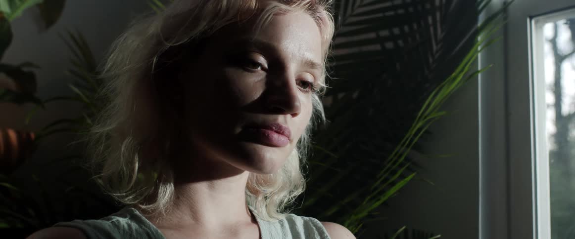 A young woman stands contemplatively next to a window in a dimly lit room during the day
