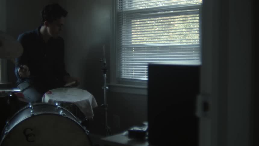 Man with a black shirt and jeans plays drums next to a window with window blind