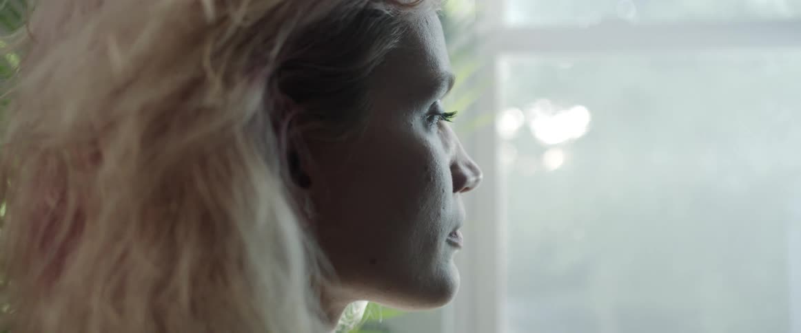 A woman with thick blonde hair stands and looks out her window on a sunny day