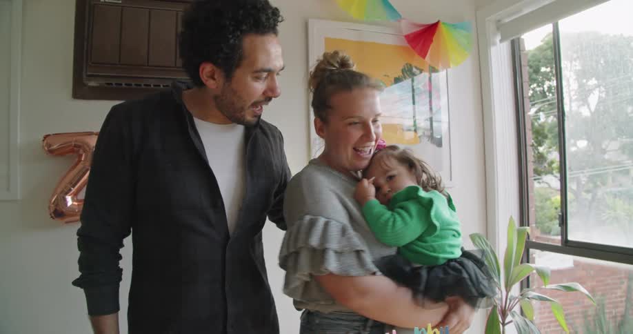 A mother and father singing happy birthday to their daughter as the mother rocks her gently from side to side standing indoors