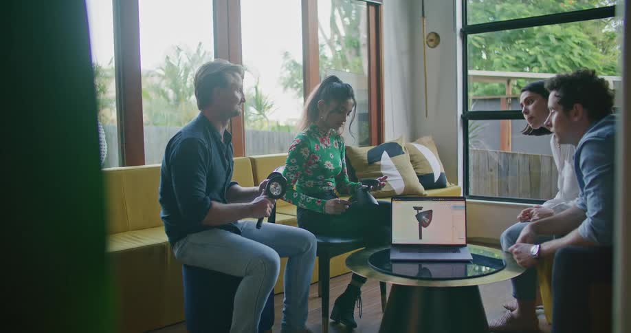 A gorup of people sit in a sun room together and pass an object back and forth with a laptop sitting on a table in between them