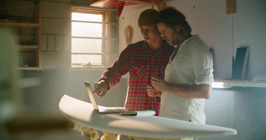 Two men discuss items on laptop screen after one examines the surfboard the laptop is sitting on
