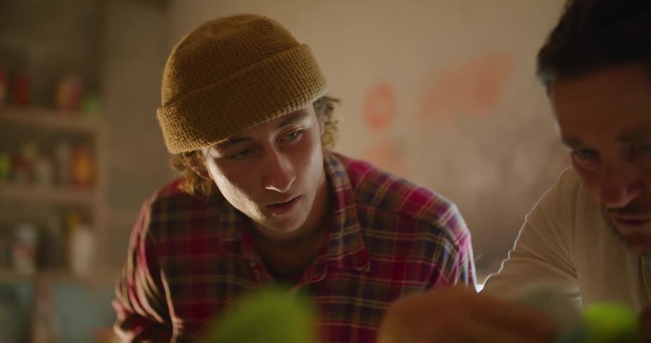 Two men in a room looking at a surfboard with a wall and window in the background