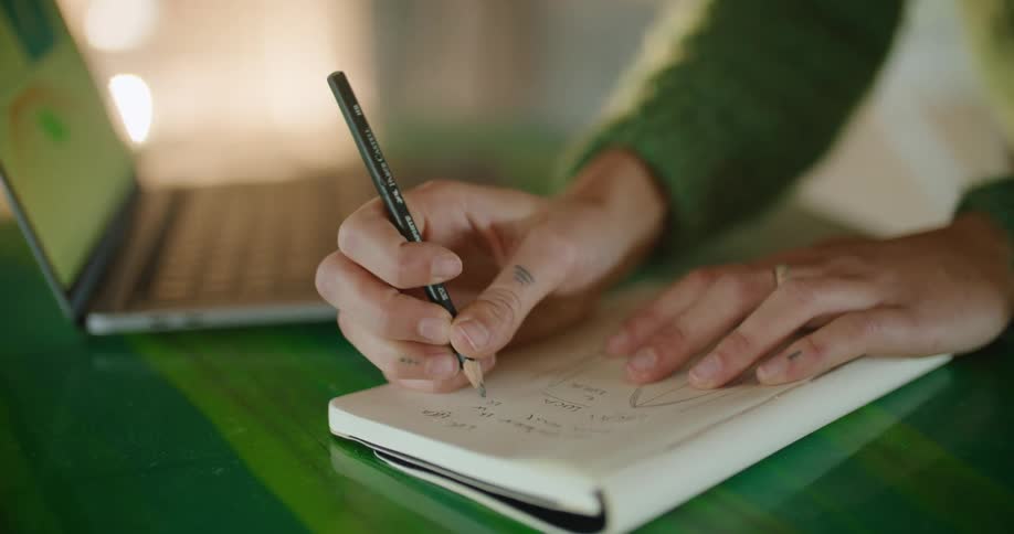 Woman with a green sweater writes in a notebook and uses a laptop