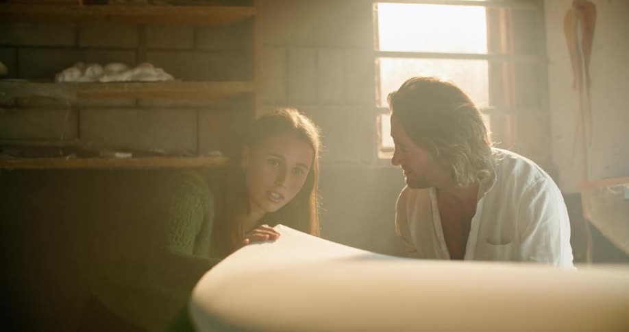 A man and woman talk and looks at a surf board being made