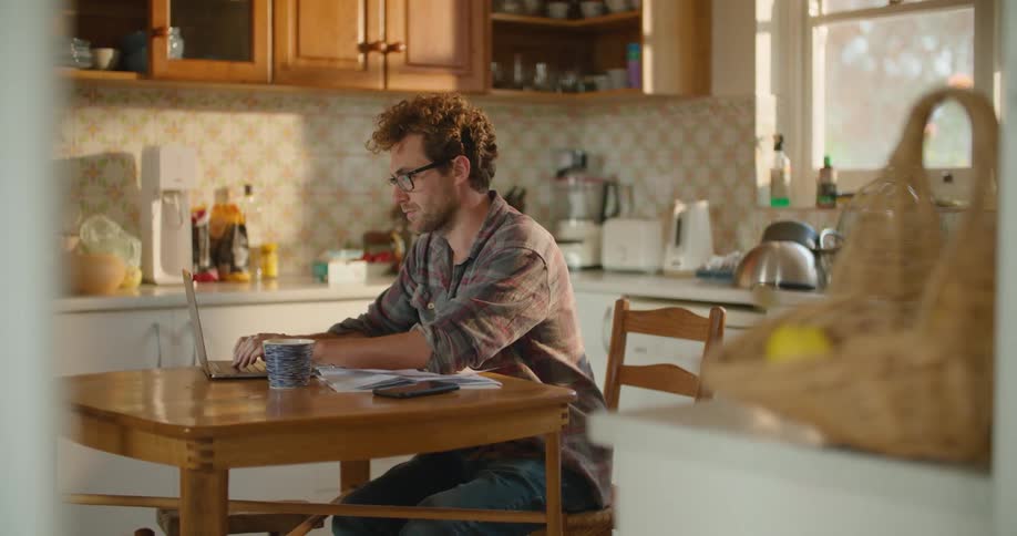 Man wearing glasses sits at a kitchen table as he types on a laptop computer