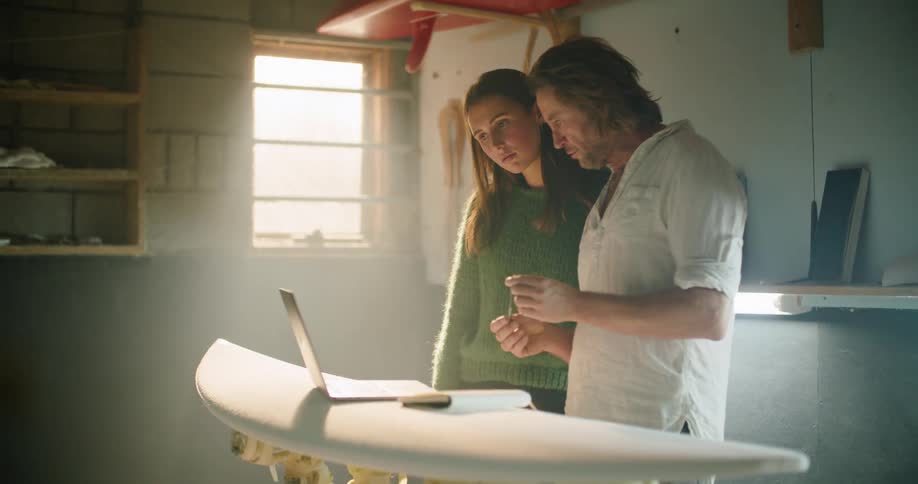 A man and woman watch video on a laptop while working on a bare surfboard
