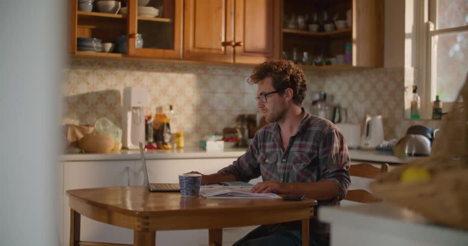 A man sits down at a small kitchen table with a mug and looks at a laptop and paper on the table