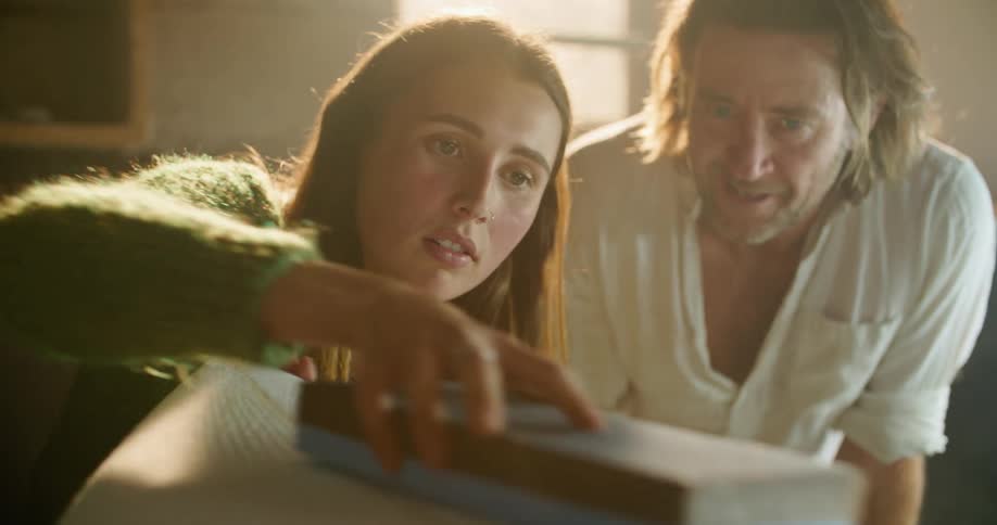 Older man teaches woman to sand surfboard properly in workshop