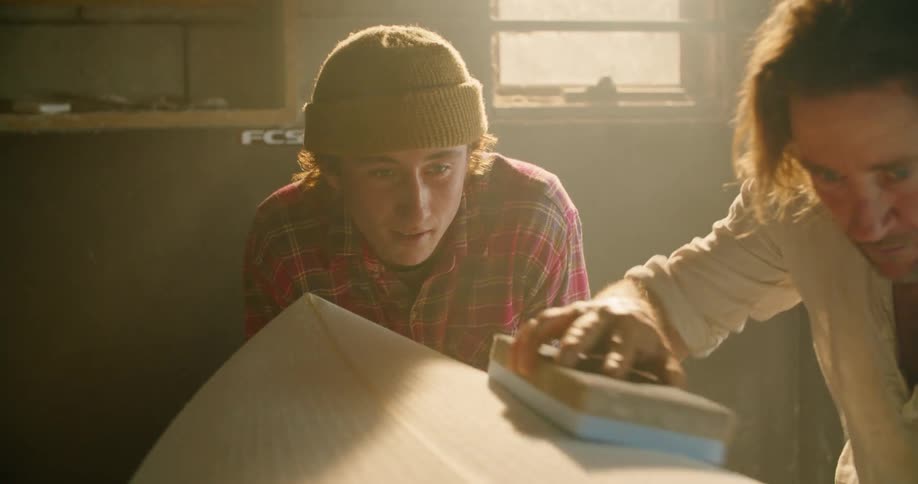 A man brushes an unfinished surfboard with a brush as a younger man watches from nearby