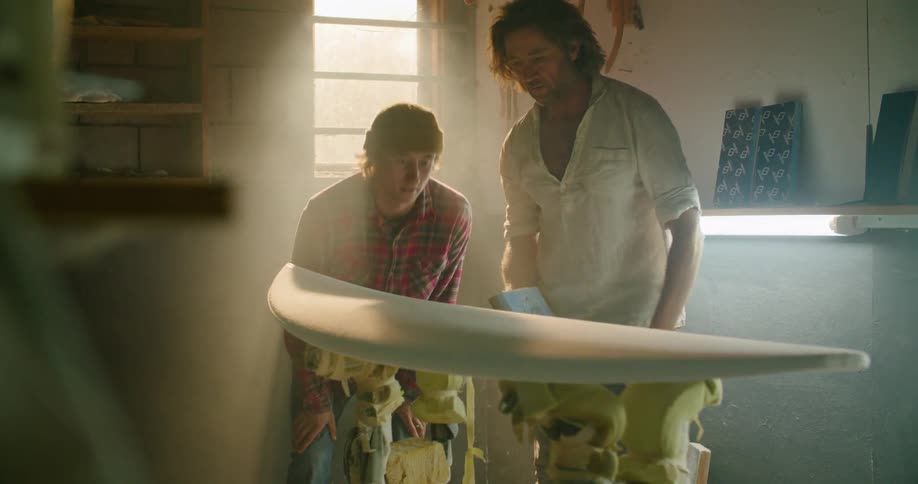 Father showing his son an unfinished surfboard as they both touch the surface inside of a small workshop