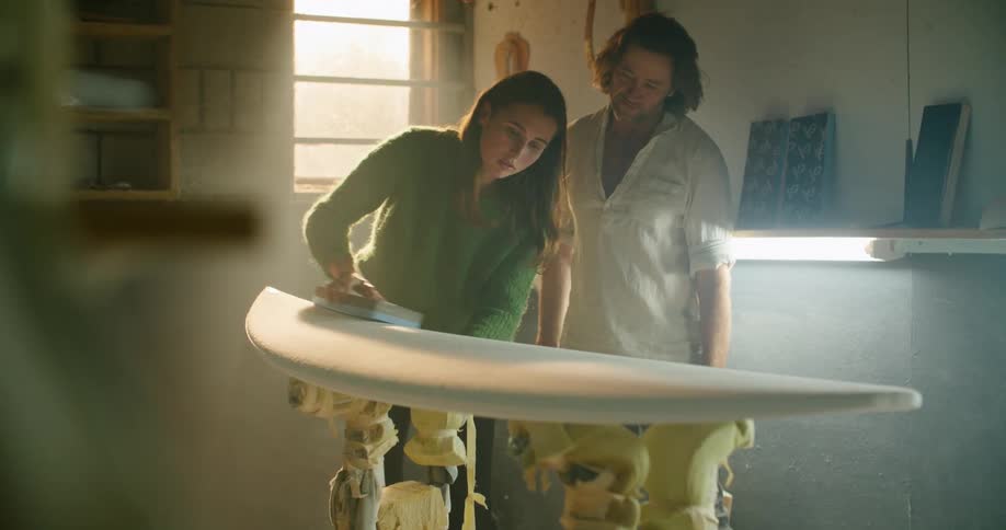 A man and a woman in a room talking as a woman sands a surfboard with a wall and window in the background