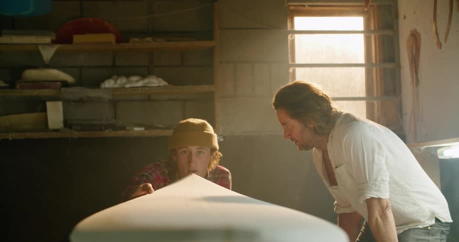 Two men in a room looking at a surfboard with a wall and window in the background