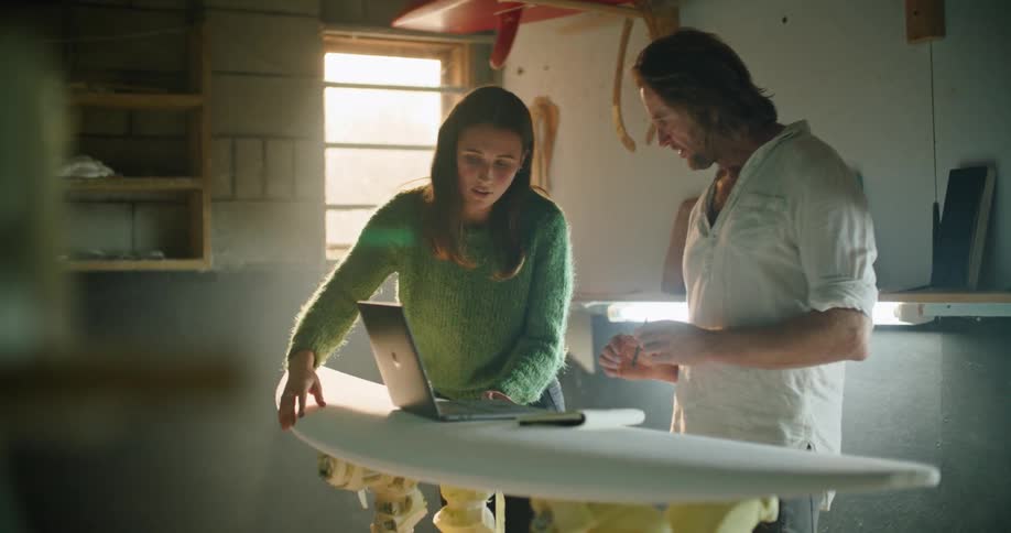 A man and a woman in a room looking at a laptop with a wall and window in the background