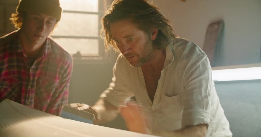 Man teaching a teenager how to sand the surface of a surfboard in a workshop