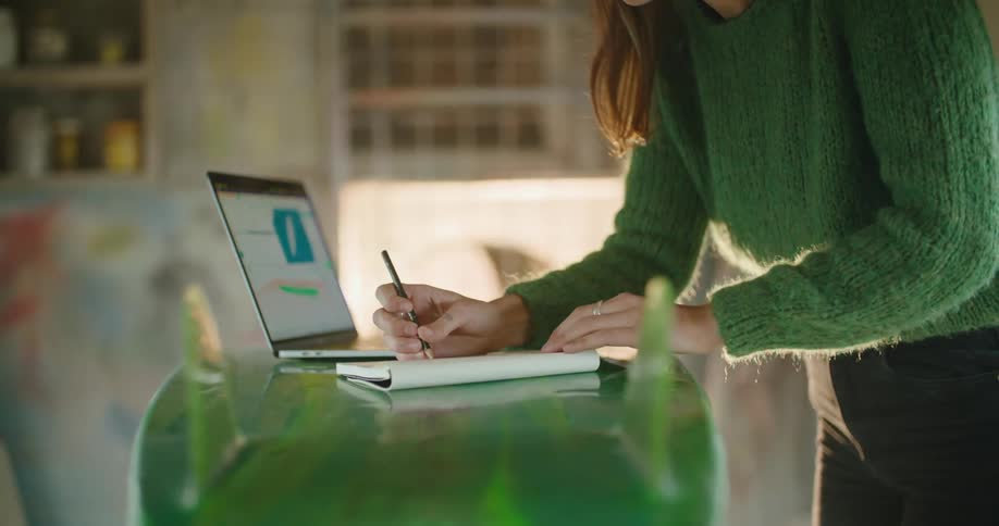 A woman works on a kitchen counter with a laptop and writes in a notebook