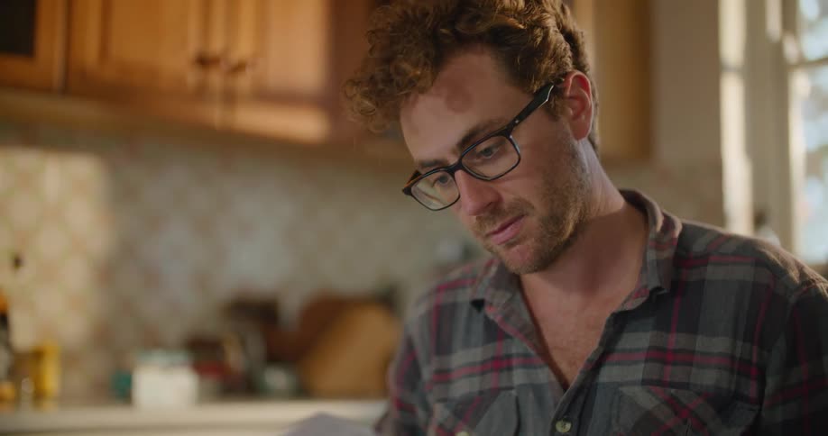 A man with glasses sitting at a kitchen table looking at papers with a wall in the background