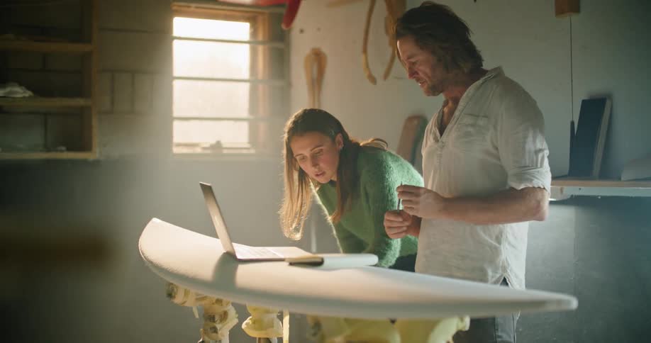 A man and woman look at a laptop and surfboard as they design it
