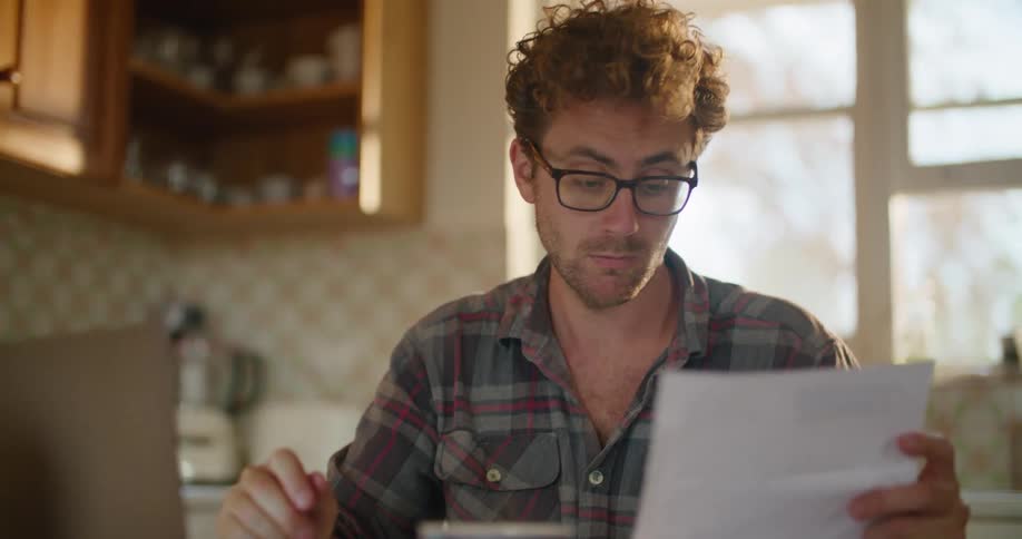 A man is sitting in his kitchen and looking at a laptop
