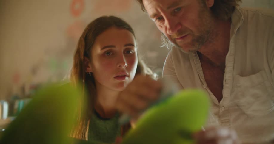 A man and a woman in a room looking at a surfboard with a wall and window in the background