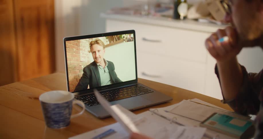 A man with glasses and a pen sitting at a table looking at a laptop