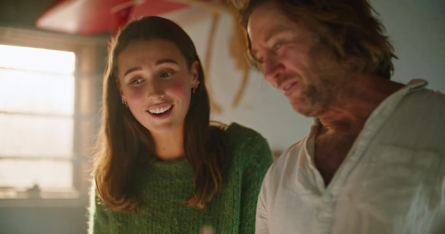 Man and woman stand in room with surfboard mounted on wall smiling and laughing as they talk