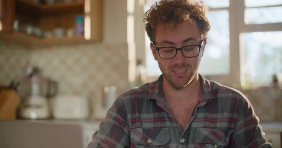 A man with glasses sitting at a table in a kitchen and looking at a laptop