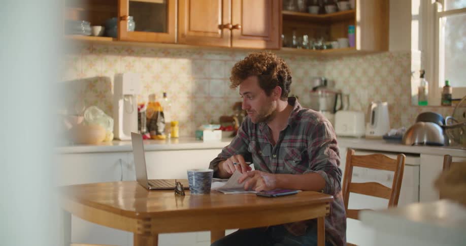 A male caucasian sitting in a kitchen at a table working on his laptop and with paperwork