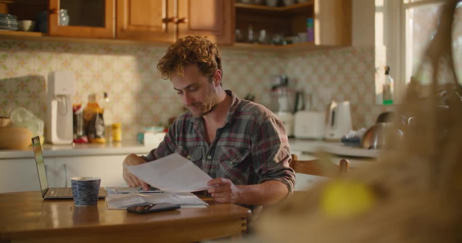 A man with glasses sitting at a kitchen table with a laptop looking at a paper