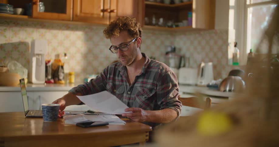 A man drinks from a coffee mug while reading a piece of paper before turning to type on a laptop as he sits at a table in a home kitchen during the day