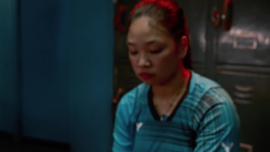 A badminton player sits in a locker room while looking down at the broken racket in their hands with a depressed expression