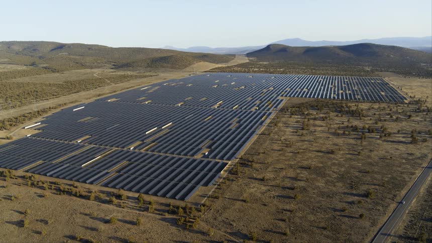 A car driving down the highway is dwarfed by a large solar farm surrounded by rolling hills and mountains on the horizon