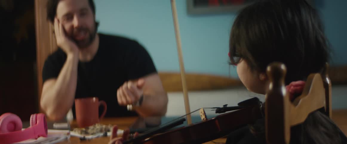 Man sits at table watching daughter practice playing musical instrument before commenting