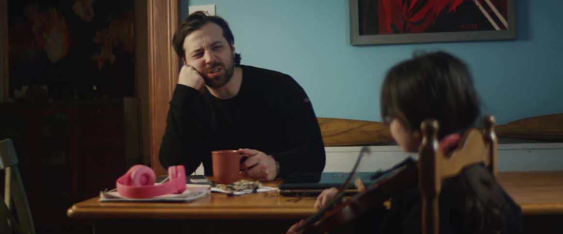A young girl with pigtails and red glasses plays the violin at a dining room table while her father encourages her in the background