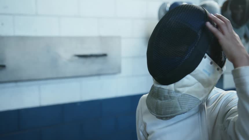 Youth standing next to tiled wall dons fencing mask