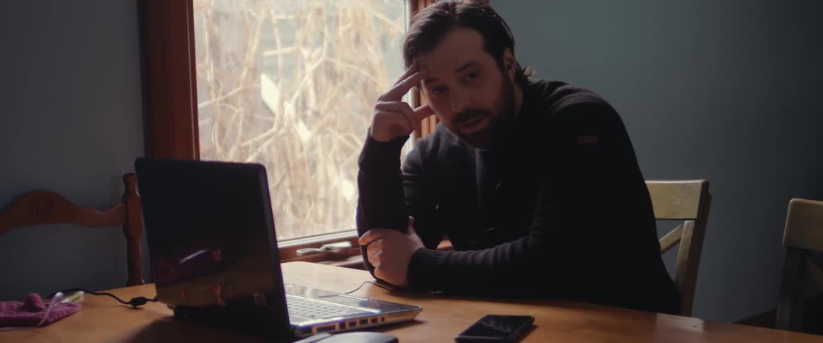 A man sitting at a table next to a small square window in front of a laptop computer with his hand on his head