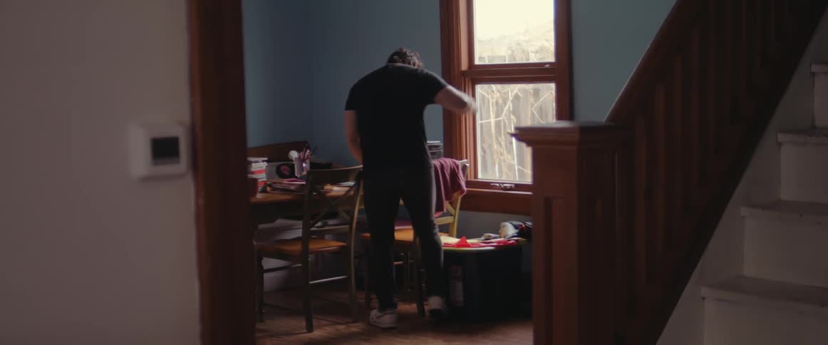 A man walks to his dining room table with a mug but starts cleaning when he sees the clutter