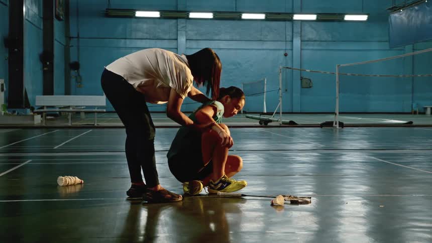 A woman walks over to a teenager sitting on the gym floor looking defeated
