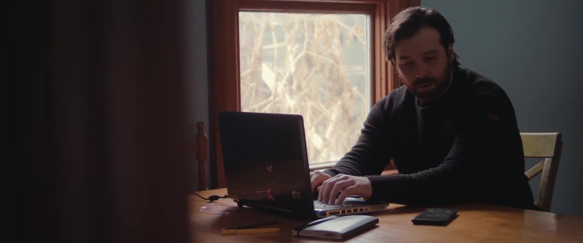 A man working from home types on his laptop and looks at his phone as it lights up and puts his head down and storms away from the table