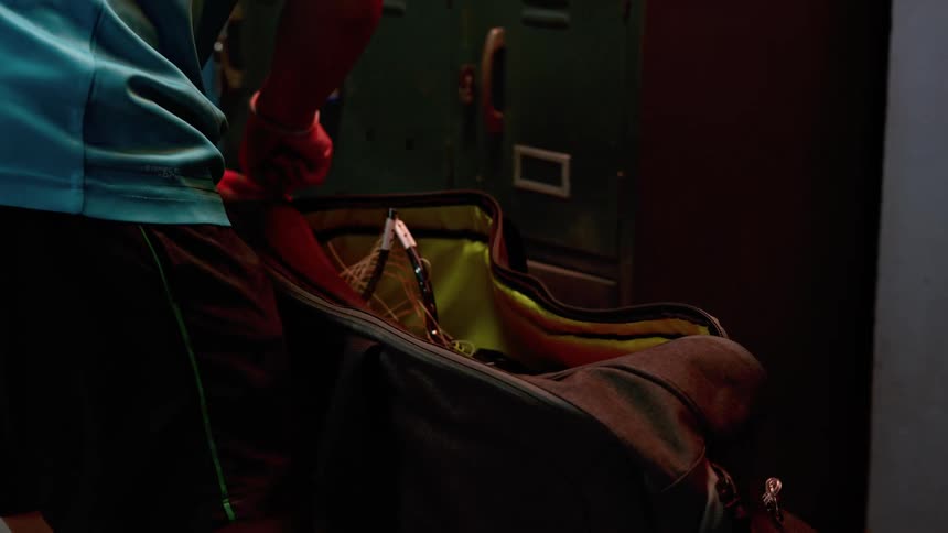 A person in a locker room putting a broken racket in a gym bag with lockers and locks in the background