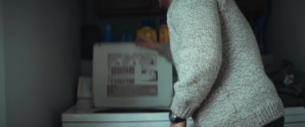 A man in a room putting clothing in a washing machine with a wall and cabinet in the background