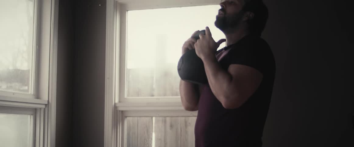 A man in a room working out with a kettlebell with a wall and window in the background