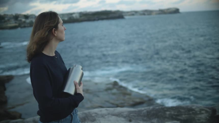 A woman holding an urn looks out contemplatively over the ocean before tossing a handful of ashes into the sea