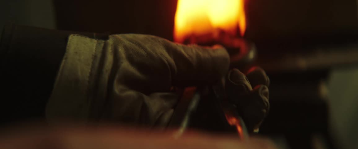 A man uses gloves and a long tool to remove a knife from a brightly burning kiln