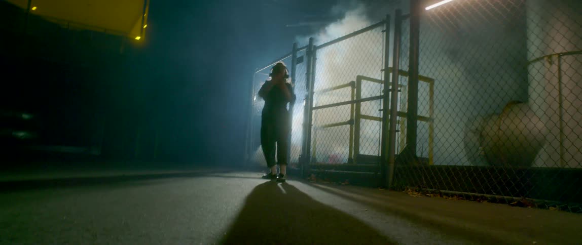 Woman dances in parking lot near chain link fence bordering factory as vapor rises from ground inside fence