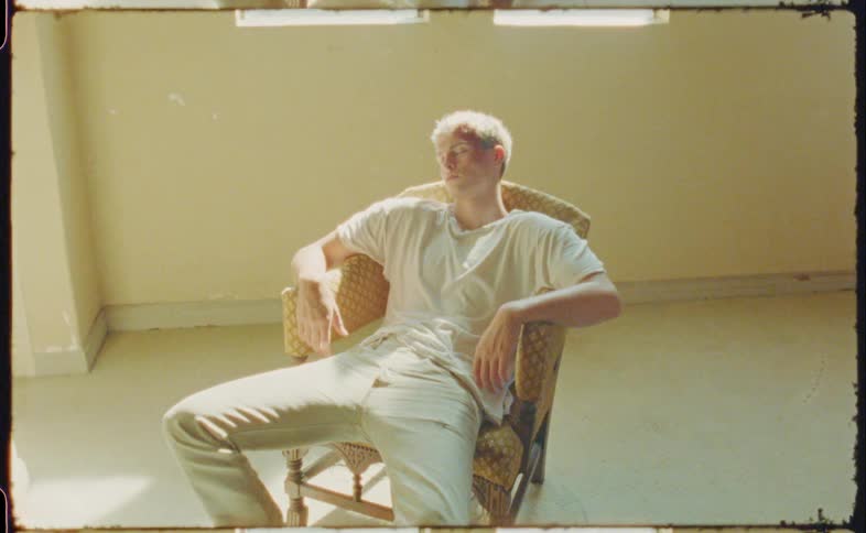 A man with blood on his head in an empty room sitting on a chair with his eyes closed