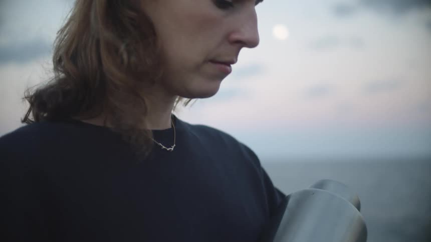A woman standing outside stares around sadly then looks down at a silver urn she is holding in her hands