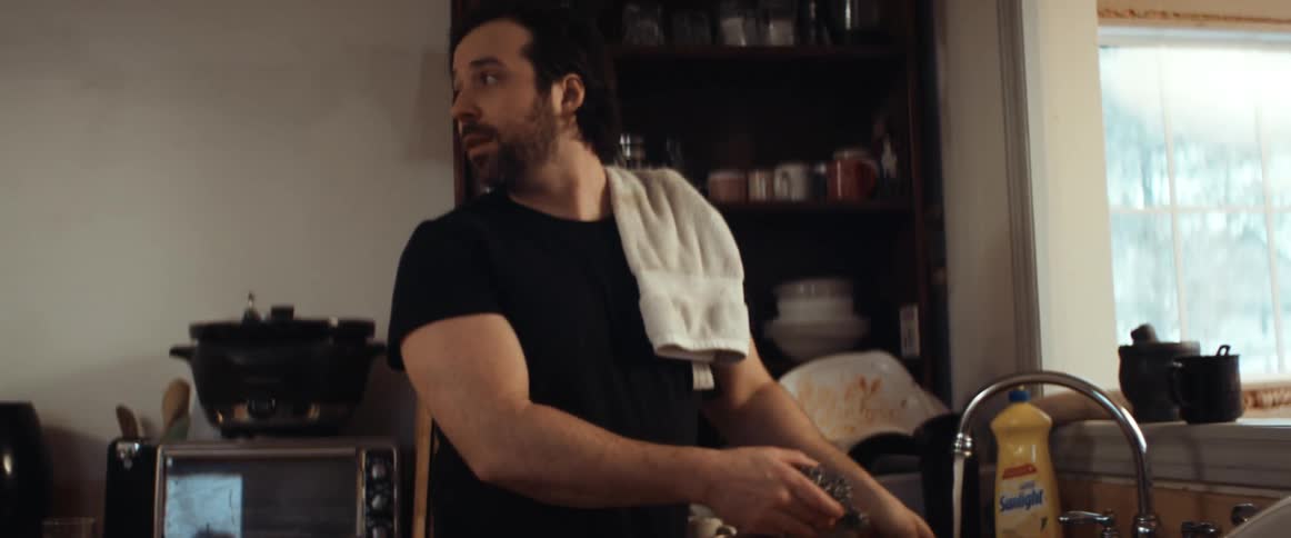 A man is doing the dishes in a home kitchen and looks behind him to talk to someone