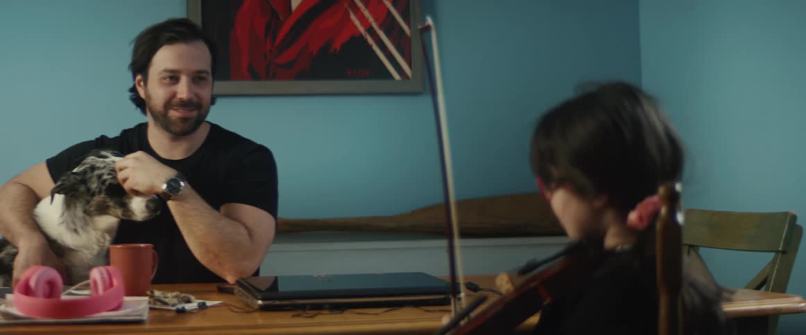 A man sits at a home table holding a dog while a girl picks up her musical instrument